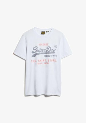 Bela bombažna majica s grafičnim potiskom v sivi in roza barvi. Oblikovanje vključuje besedilo "Vintage SuperDry" in akcenta iz Tokia, Japonska.