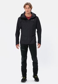 Giacca nera impermeabile con cappuccio regolabile, chiusura zip frontale e due tasche sul petto. Abbinata a pantaloni neri con dettagli testurizzati e riflettenti.