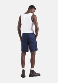 Pantalones cortos azul marino con dos bolsillos traseros, combinados con una camiseta sin mangas blanca y botines marrones hasta el tobillo. La tela parece ligera y casual.