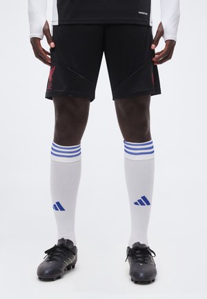 Athlète masculin portant un short de football Liverpool noir, des chaussettes montantes blanches Adidas avec des rayures bleues, et des crampons noirs, debout devant un fond blanc.
