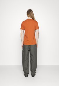 Krátký rukáv oranžového trička a volné šedé cargo kalhoty s bočními kapsami, nošené s černými botami, pohled zezadu.