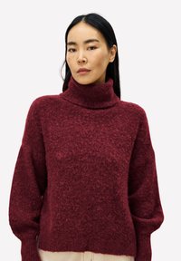 Frau mit langen, dunklen Haaren, die einen strukturierten burgunderfarbenen Rollkragenpullover vor einem schlichten hellen Hintergrund trägt.