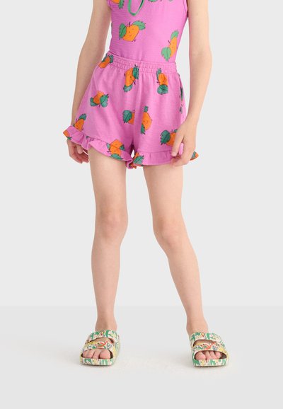Enfant portant un short violet et un haut avec un imprimé de fruit orange et des bords volantés, associé à des sandales ouvertes à motifs sur fond blanc.