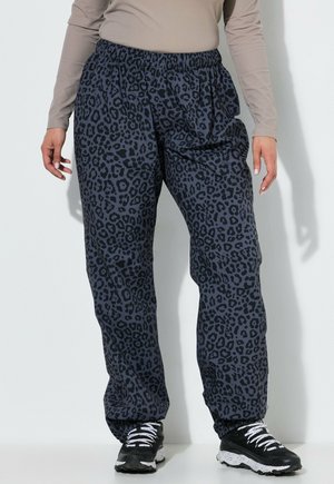 Person iført løstsiddende mørkegrå leopardmønstrede bukser, sort-hvide sneakers og en beige langærmet top, stående op ad en hvid væg.