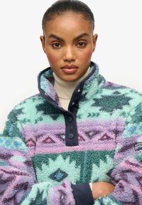 Pull polaire en vert, violet et rose avec des motifs géométriques, col montant et boutons ; possède une surface douce et texturée.