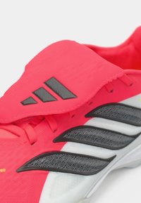 Ροζ αθλητικό παπούτσι με υφή στην επάνω πλευρά, διαθέτει μαύρες πλευρικές ρίγες και ιμάντα με βέλκρο. Η σόλα είναι λευκή με λεπτομέρειες.