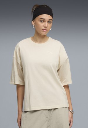 Beige oversized T-shirt met korte mouwen, zachte katoenen stof, ronde hals en subtiel logo op de borst. Soepele textuur en een ontspannen pasvorm.