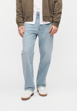 Light blue denim jeans with a relaxed fit, featuring five pockets and a classic button fly. Paired with white sneakers and a green zip-up hoodie.