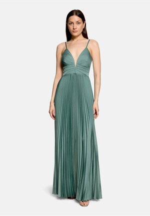 Femme portant une robe maxi verte plissée sans manches avec un profond décolleté en V et de fines bretelles, debout devant un fond clair uni.