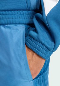 Chaqueta deportiva azul con un tejido texturizado, puños acanalados y bolsillos con un forro suave. Se aprecian acentos blancos en contraste.