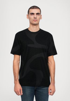 T-shirt noir à manches courtes avec un motif texturé affichant un texte stylisé. Encolure ronde et coupe décontractée. Porté avec un jean bleu.