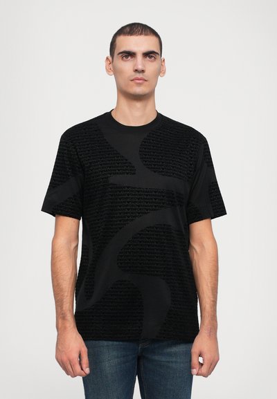 Czarny T-shirt z krótkim rękawem o teksturowanym wzorze z stylizowanym napisem. Okrągły dekolt i luźny krój. Noszony z niebieskimi dżinsami.