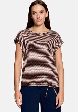 Femme aux cheveux bruns et raides porte un haut rayé à manches courtes avec un détail noué sur le bas et un pantalon bleu foncé.