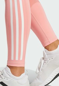 Różowe legginsy sportowe mają trzy pionowe białe paski wzdłuż zewnętrznej części uda. Materiał jest gładki i elastyczny, w połączeniu z białymi butami sportowymi.