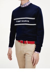 Man med marinblå tröja med texten "TOMMY HILFIGER", röd flätad bälte, beige byxor och rutiga skjortärmar synliga vid handlederna och kragen.