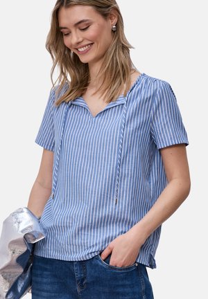 Uśmiechnięta kobieta w krótkim rękawie, niebiesko-białej pasiastym topie i niebieskich dżinsach, trzymająca w lewej ręce błyszczący srebrny materiał.