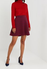 Maglione a collo alto rosso abbinato a una gonna a ruota a strisce blu navy e bordeaux. L'outfit presenta una silhouette aderente e un tessuto strutturato.