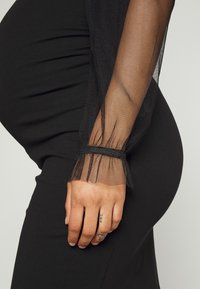 Vestido preto com uma silhueta ajustada e mangas longas em malha. As extremidades das mangas têm um acabamento com babados e são franzidas no pulso.