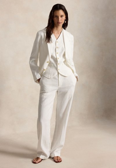 Γυναίκα που φοράει ένα λευκό ραμμένο κοστούμι με σακάκι, γιλέκο με κουμπιά, παντελόνια φαρδιάς γραμμής και σανδάλια, στέκεται μπροστά σε ένα απλό μπεζ φόντο.