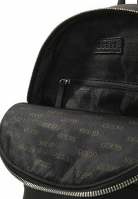 Black backpack interior features a smooth fabric lining with repeating "GUESS" logo pattern, including a zippered compartment and durable zipper.