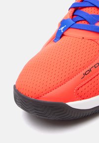 Κόκκινο αθλητικό παπούτσι με υφασμάτινο πάνω μέρος από ανάγλυφο πλέγμα, μπλε κορδόνια και μαύρη καουτσούκ σόλα με μοτίβο ψαροκόκαλο; λεπτομέρεια λογότυπου στην γλώσσα.