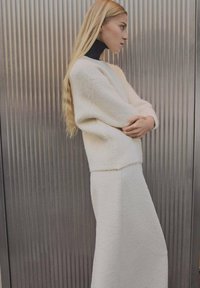 Pull blanc duveteux et jupe midi assortie, présentant une finition texturée. Le mannequin se tient contre un fond à côtes verticales.