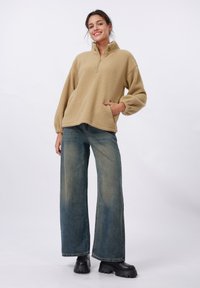 Beige fleece pullover met een halfrits en brede mouwen, gecombineerd met vervaagde blauwe wijde jeans en zwarte platformlaarzen.