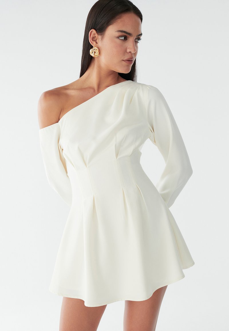 Abito bianco asimmetrico con maniche lunghe, corpino attillato e gonna svasata. Tessuto liscio con pieghe laterali e dettagli minimi.