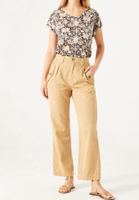 Květinový vzorovaný černý top s krátkými rukávy, spárovaný s béžovými kalhotami s širokými nohavicemi, které mají kapsy a detaily na knoflíky. Hnědé sandály na řemínky.