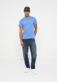 Plava majica s okruglim izrezom i natpisom "tommy" u crvenoj i bijeloj boji, u kombinaciji s tamnoplavim trapericama i bijelim cipelicama. Crna kapa upotpunjuje izgled.