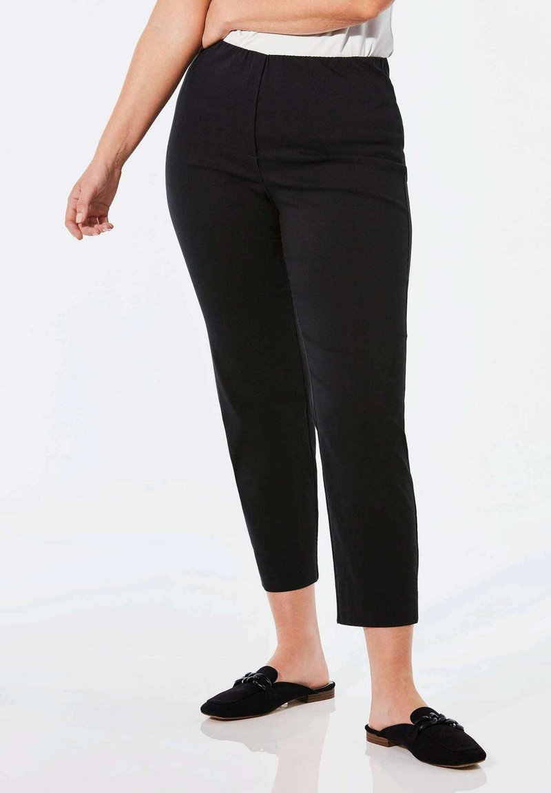 Pantalon noir ajusté avec une taille élastique et une longueur de jambe raccourcie, associé à des chaussures plates noires à enfiler avec des accents en forme de nœud.