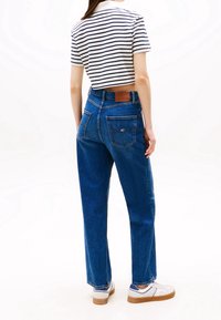 Osoba stojící před bílým pozadím, oblečená v modrých džínech s rovnými nohavicemi, bílých teniskách a bílém krátkém tričku s černými horizontálními pruhy.