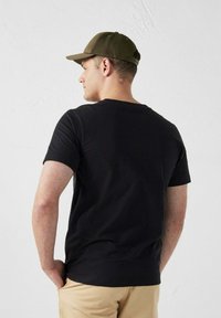 Zwarte t-shirt met korte mouwen, ronde halslijn, gedragen met kaki shorts. Een donkergroene pet wordt achterstevoren gedragen. Eenvoudig ontwerp, casual pasvorm.