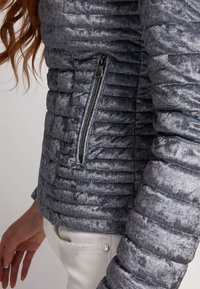Gros plan sur une personne portant une veste matelassée grise texturée avec une poche zippée sur le côté et un pantalon blanc, montrant une partie de son bras et de sa main.