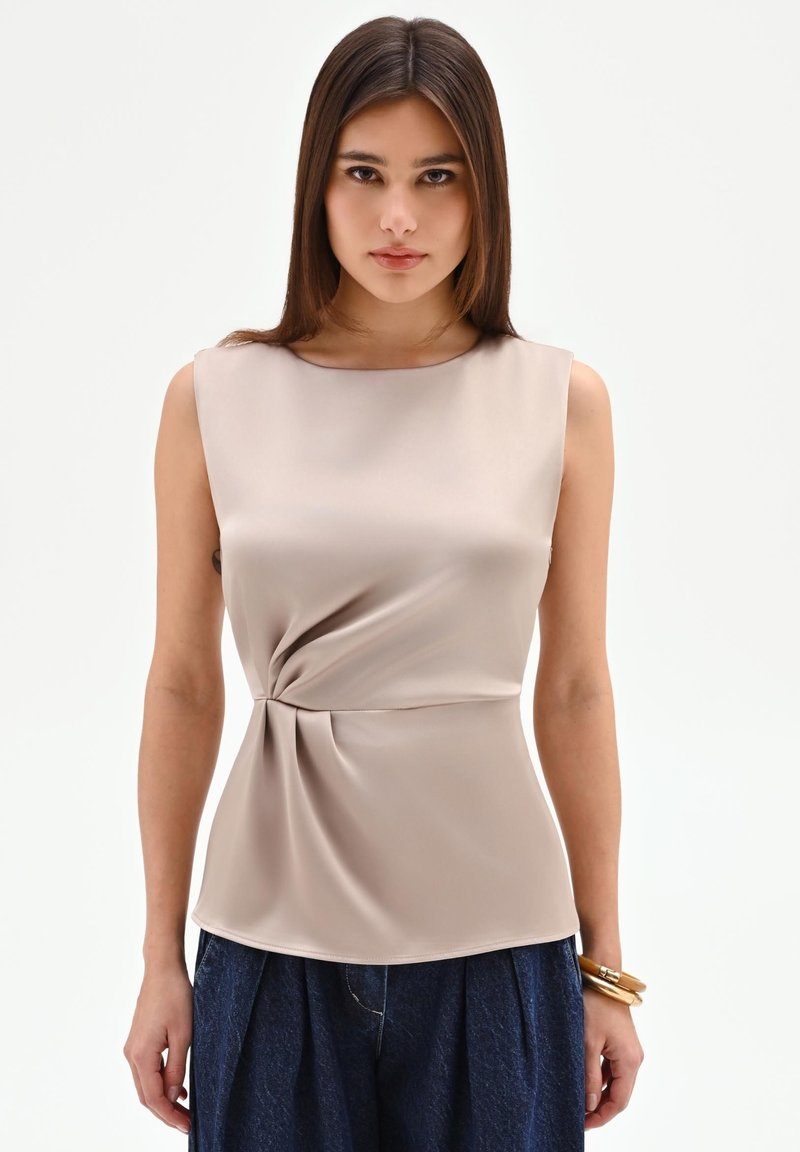 Jonge vrouw met recht bruin haar draagt een mouwloze beige satijnen top met zijwaartse twist en donkerblauwe geplooide broek, gouden armband.