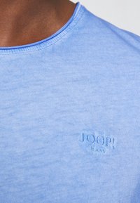 Světle modré bavlněné tričko s kulatým výstřihem, na kterém je jemně vyšívané logo "JOOP! JEANS" v mírně tmavší modré barvě.