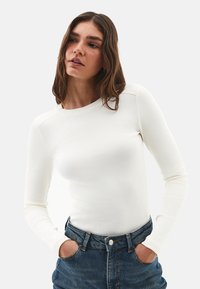 Giovane donna con capelli castani ondulati che indossa un top fitted bianco a coste a maniche lunghe e jeans di denim blu, con le mani nelle tasche, che guarda di lato.