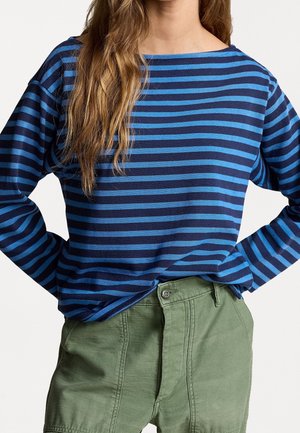 Person trägt blau- und marineblau gestreiftes Langarmshirt und grüne Hose, Hände in die Hüften gestemmt, Haare fallen über eine Schulter.