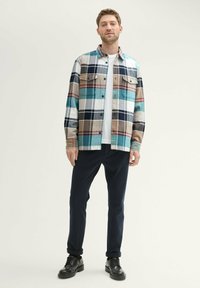 Flanellhemd im mehrfarbigen karrierten Muster (blau, beige und braun) mit Knopfleiste an der Vorderseite, zwei Brusttaschen, getragen über einem weißen T-Shirt und schwarzen Hosen.