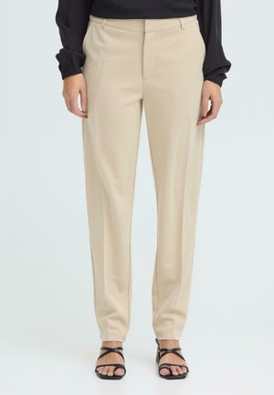 Kobieta ubrana w beżowe spodnie o prostych nogawkach, czarną bluzkę z długim rękawem i czarne sandały na obcasie z odkrytymi palcami, stojąca na jednolitym tle.