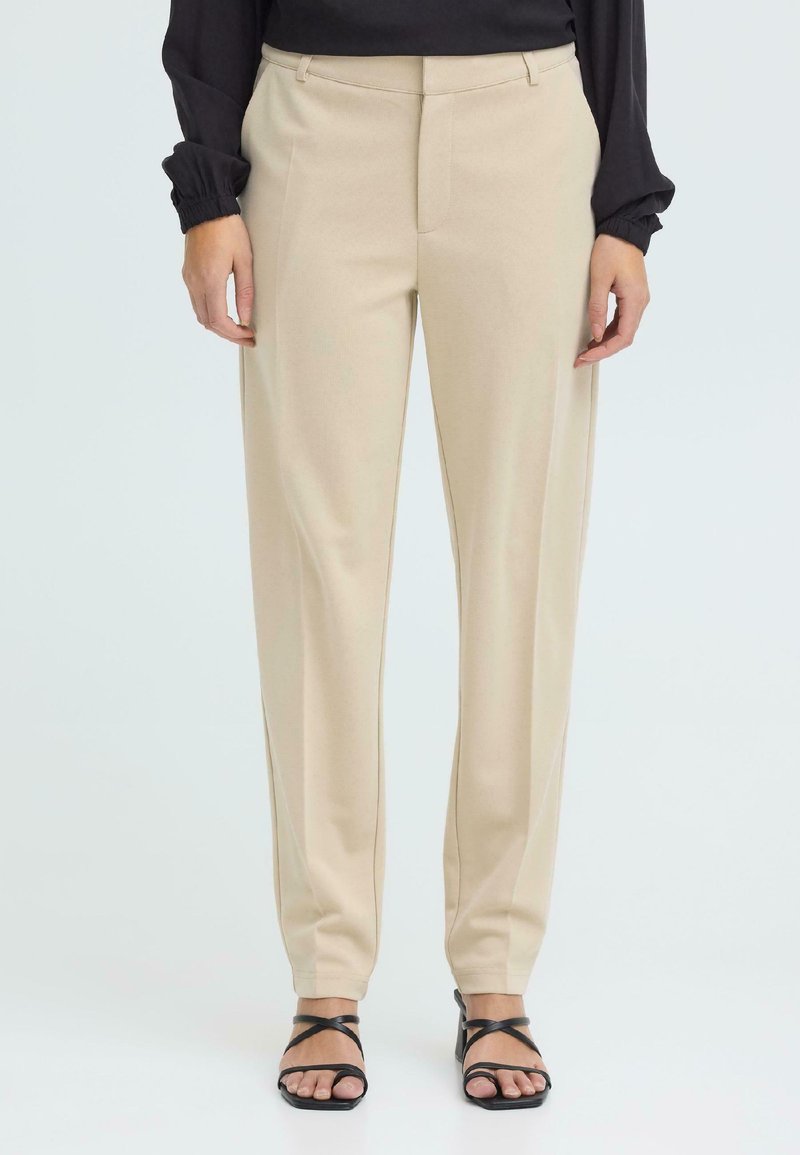 Femme portant un pantalon beige à jambes droites, un haut noir à manches longues et des sandales à talons noires ouvertes, posant devant un fond uni.