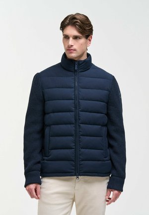 Uomo che indossa una giacca trapuntata blu navy con maniche lavorate a maglia e pantaloni beige, in piedi davanti a uno sfondo chiaro e uniforme.