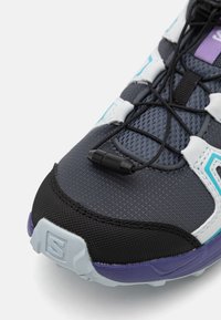 Pelēki sporta apavi ar melniem akcentiem, strukturētu sietu augšdaļu, elastīgu šņorēšanas sistēmu un purpura detaļām. Ievērojama izliekta zole un logo uz papēža.