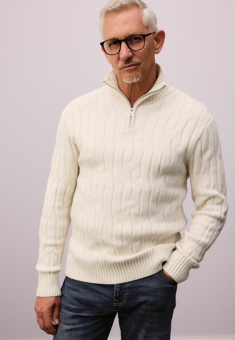 Suéter de punto trenzado color crema con cuello de media cremallera, puños y dobladillo acanalados, combinado con jeans de mezclilla. Textura suave y detalle de patrón sutil.