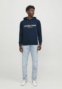 Sudadera marina con un bolsillo frontal, que presenta texto de logo en blanco. Combinada con jeans de color azul claro y zapatillas negra y blanca.