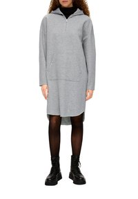 Šedé šaty s kapucí na zip a přední kapsou, dlouhými rukávy a bočními rozparky; v kombinaci s černými kotníkovými botami a neprůhlednými punčocháči.