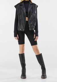 Chaleco sin mangas de cuero sintético negro con solapas en los hombros, combinado con una camiseta ajustada de manga larga negra y pantalones cortos de ciclista de cintura alta. Botas de plataforma negras hasta la rodilla.