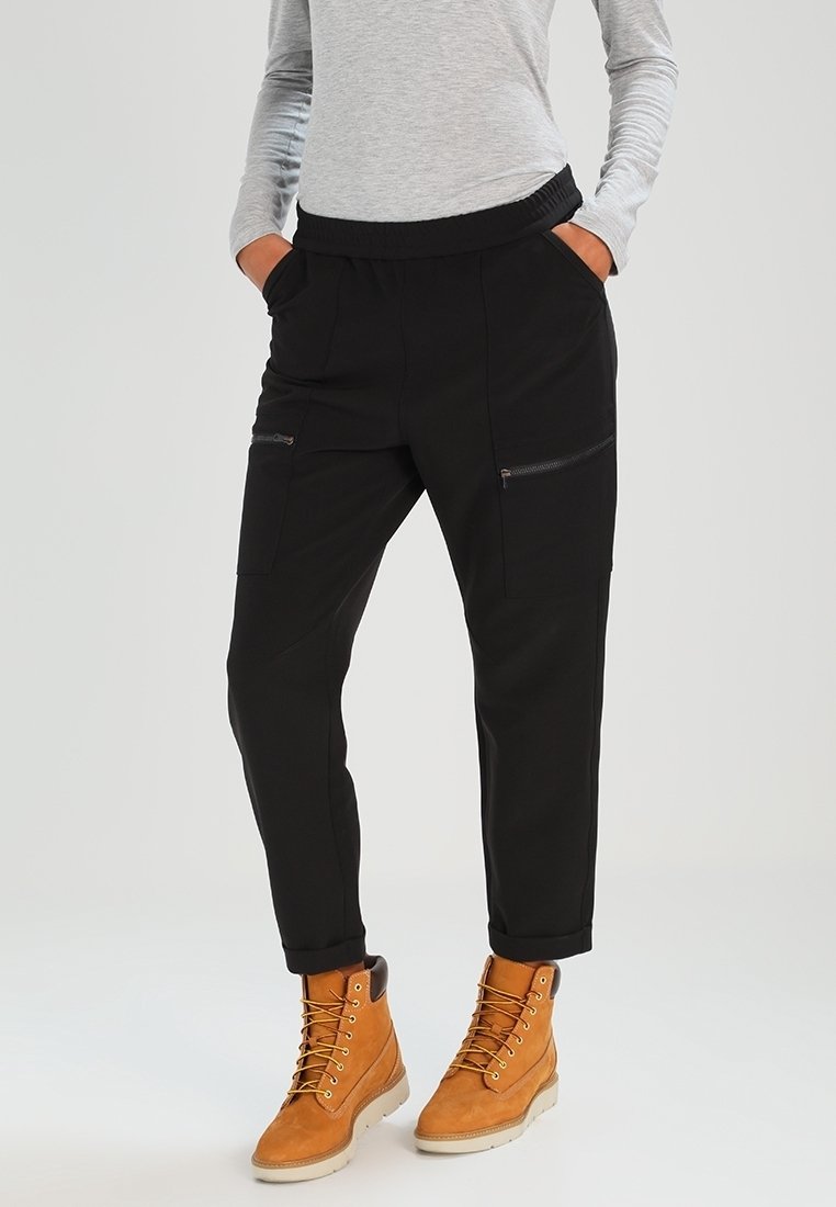 Pantalon cargo noir avec taille élastique, poches avant et détails zippés, associé à des bottes marron clair avec lacets et semelles en caoutchouc.