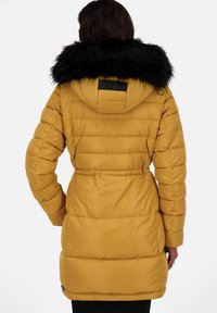 Vrouw in een mosterdgeel gewatteerd jasje met een capuchon van zwart nepbont, gezien van achteren tegen een effen witte achtergrond.