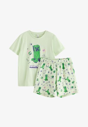 Garçon souriant en pyjama vert clair thème Minecraft avec des motifs Creeper et des chaussons verts Creeper assortis, debout sur fond blanc.